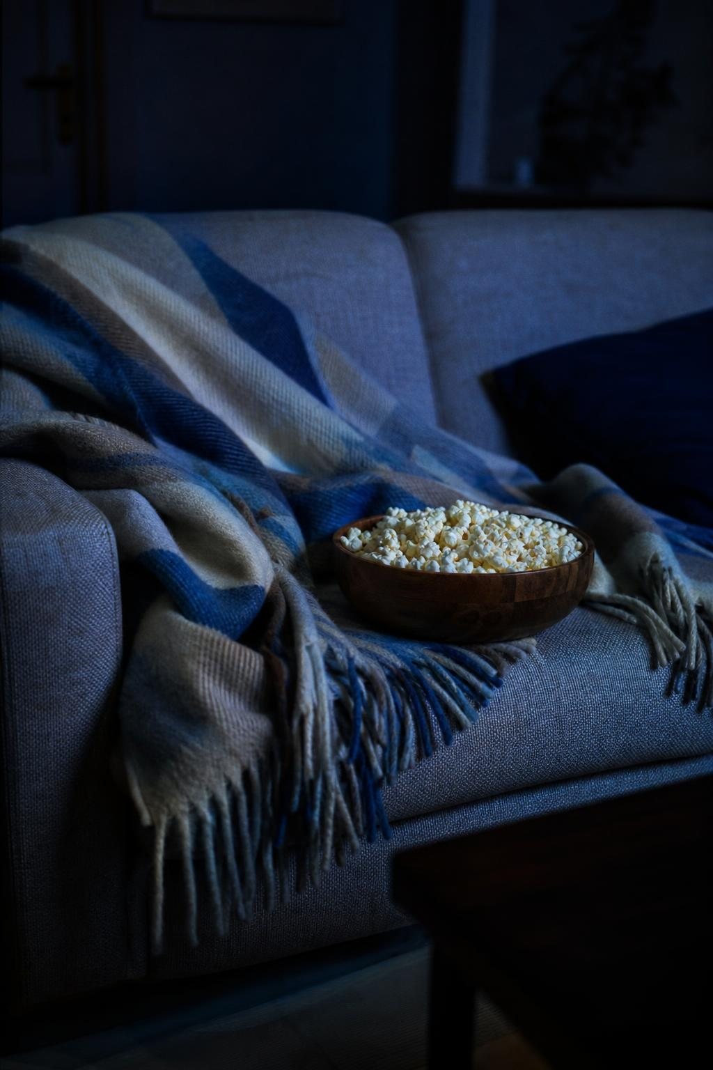 Sofa mit Wolldecke und Popcornschale im dunklen Wohnzimmer, nur vom bläulichen Licht eines Fernsehers beleuchtet (mit AI erstelltes Bild)