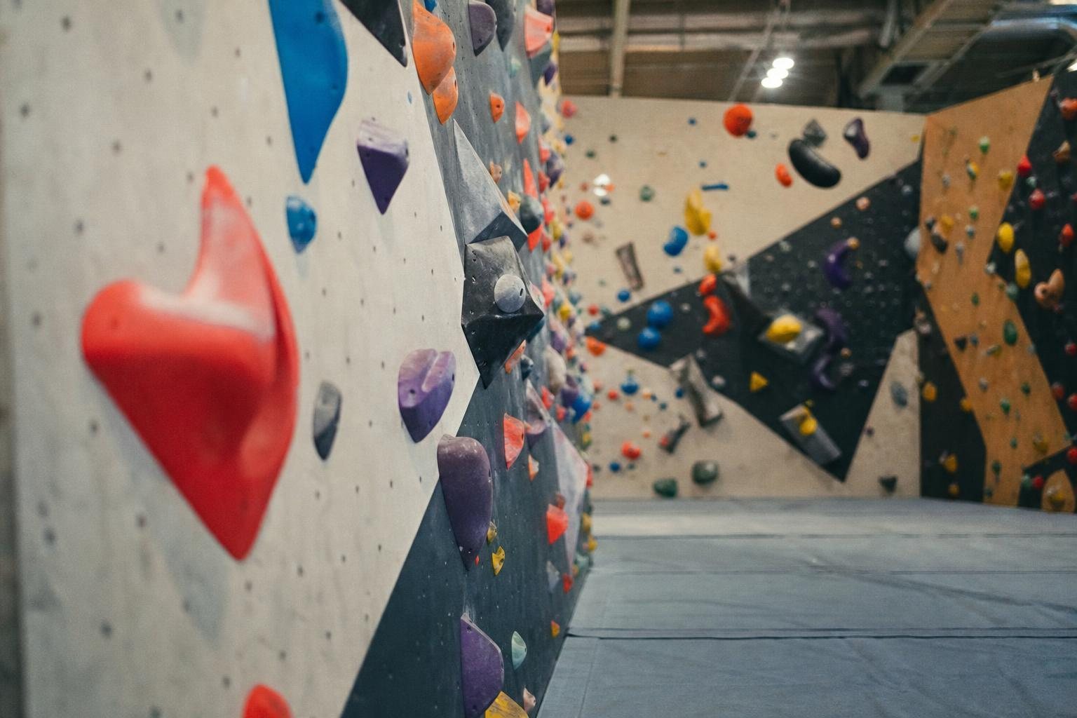Boulderwand mit bunten Klettergriffen und Fallschutzmatten in einer hellen leeren Halle (mit AI erstelltes Bild)